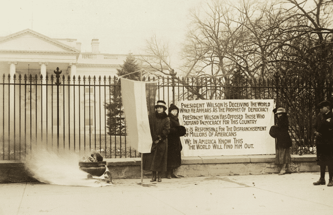 Photograph of National Woman's Party watchfire demonstrators standing with banners and fire in urn in front of White House. One banner reads, "President Wilson is deceiving the world when he appears as the prophet of democracy. President Wilson has opposed those who demand democracy for this country. He is responsible for the disfranchisement of millions of Americans. We in America know this. The world will find him out."