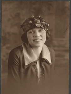 Half-length portrait of Nell Mercer, seated, with painted backdrop, wearing broad-collared suit and hat with leaves and berries. Printed as a picture postcard.
