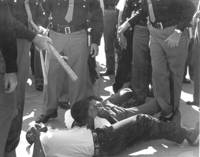 While FBI agents stood by, SNCC volunteers were beaten and arrested by sheriff deputies for attempting to bring water to people (many elderly) waiting in the hot sun for hours to register to vote. © John Kouns.
