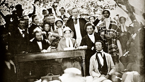The 1850 Fugitive Slave Law Convention, Cazenovia, New York. The Edmonson sisters are standing wearing bonnets and shawls in the row behind the seated speakers. Frederick Douglass is seated, with Gerritt Smith standing behind him, and with Abby Kelley Foster the likely person seated on Douglass's left. Image: WikiCommons.