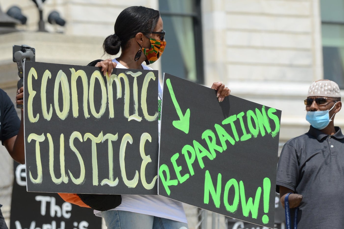 protester holding up signs reading "Economic Justice" and "Reparations Now!"